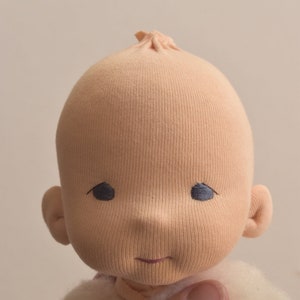 May include: A close-up of a handmade cloth doll's face. The doll has a light peach-colored face with embroidered eyes and a small smile. The doll's head is made of a soft, textured fabric.