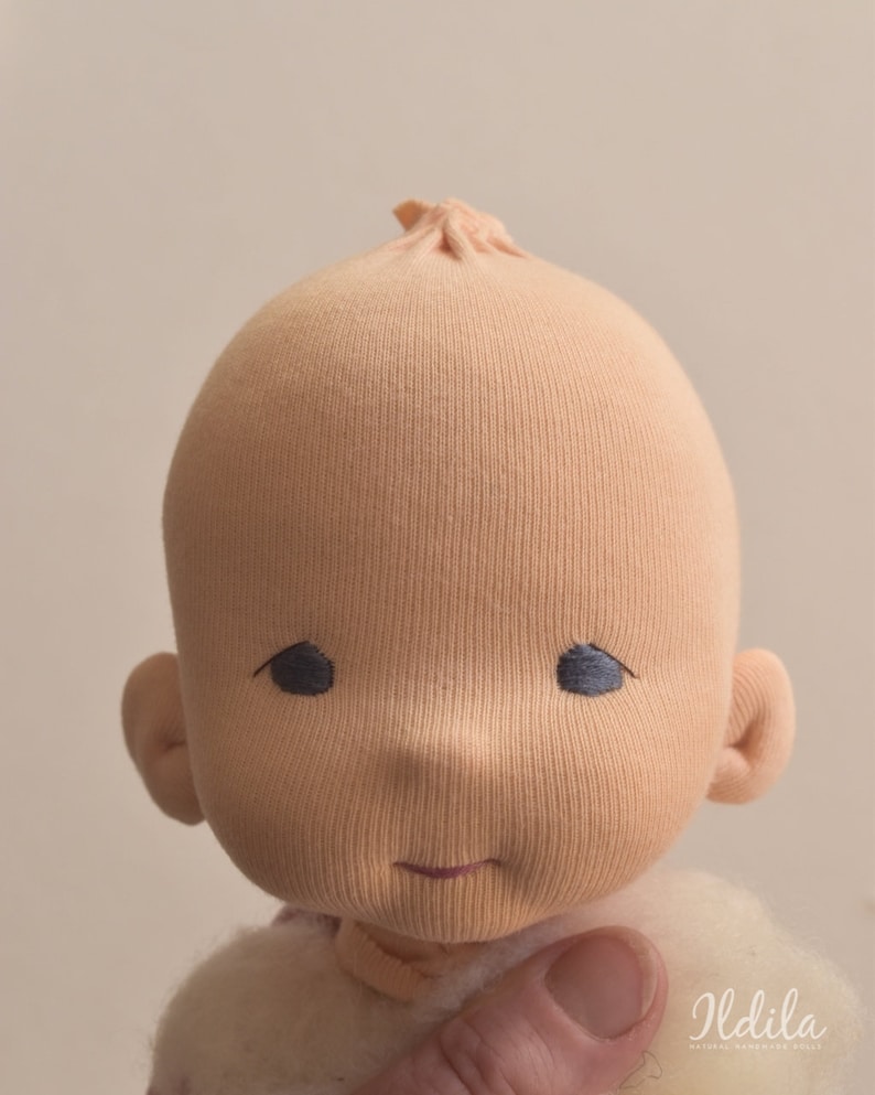 May include: A close-up of a handmade cloth doll's face. The doll has a light peach-colored face with embroidered eyes and a small smile. The doll's head is made of a soft, textured fabric.