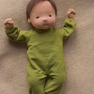 May include: A handmade cloth doll with brown hair wearing a green jumpsuit. The doll is lying on a light brown fabric surface.