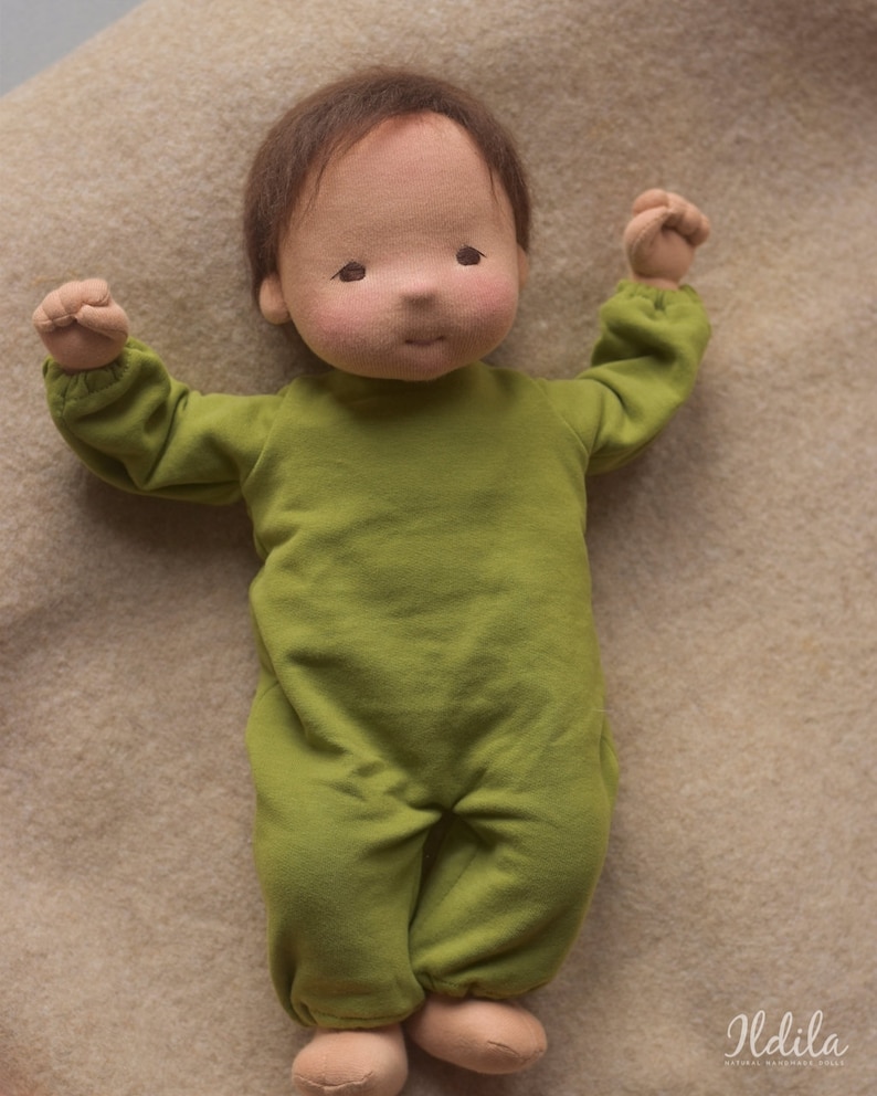 May include: A handmade cloth doll with brown hair wearing a green jumpsuit. The doll is lying on a light brown fabric surface.