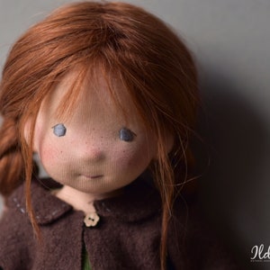 May include: Close-up of a handmade doll with red hair, blue eyes, and a brown sweater. The doll has a sweet expression and is made with natural materials.