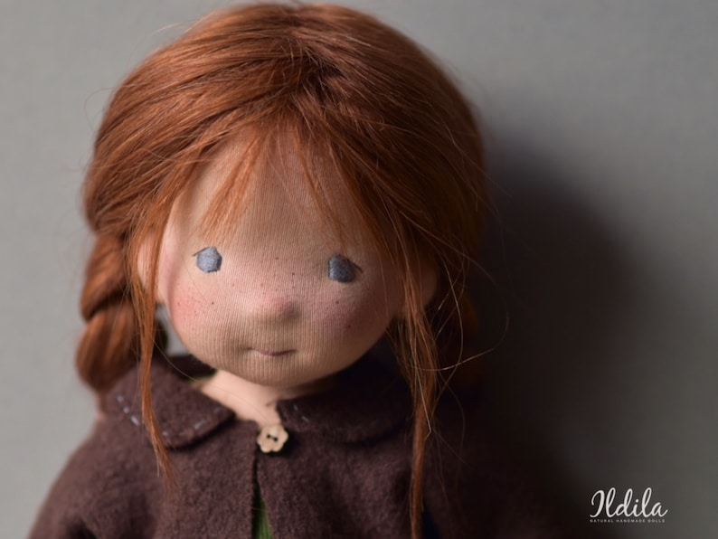 May include: Close-up of a handmade doll with red hair, blue eyes, and a brown sweater. The doll has a sweet expression and is made with natural materials.
