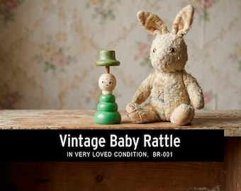 May include: A vintage baby rattle and a stuffed rabbit are displayed on a wooden surface. The rattle is green and wooden, with a smiling face and a hat. The rabbit is beige with long ears and a worn appearance. Text on the image reads "Vintage Baby Rattle".