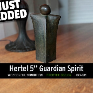 Puede incluir: Una escultura Hertel Guardian Spirit de color bronce, de aproximadamente 13 cm de altura, con una parte superior redondeada y un cuerpo rectangular. La escultura está sobre una superficie de madera. El texto "JUST ADDED" está en la esquina superior izquierda.