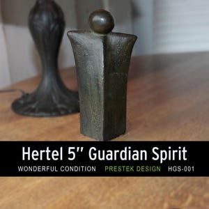 Puede incluir: Una escultura de color bronce, etiquetada como "Hertel 5" Guardian Spirit", se encuentra sobre una superficie de madera. La escultura mide aproximadamente 13 cm de altura y tiene una forma humana estilizada. La base es rectangular y la parte superior tiene una cabeza redondeada. También se ven las palabras "Wonderful Condition".
