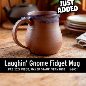 May include: A brown ceramic mug with a purple glazed rim and a small blue bead attached to the handle. The text on the image reads: JUST ADDED, Laughin' Gnome Fidget Mug, PRE 2024 PIECE, MAKER STAMP, VERY NICE, LG001. The mug has a rounded body shape.