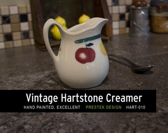 May include: A vintage Hartstone creamer, hand-painted with fruit designs. The white creamer has a handle and spout. The text "Vintage Hartstone Creamer" is displayed below the creamer. The creamer is a kitchenware item.