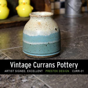 May include: A vintage ceramic vase from Currans Pottery, featuring a speckled off-white and blue glaze. The vase has a rounded form and a narrow opening. The text "Vintage Currans Pottery" is displayed on a black banner.