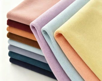 May include: A stack of folded fabric pieces in various solid colors including pale yellow, peach, light blue, lavender, dusty rose, burgundy, orange, brown, light gray, teal, and dark navy blue. Each piece features a fine, ribbed knit texture.