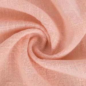May include: A soft, light peach-colored fabric with a visible woven texture. The material is gathered into a central swirl, creating deep, flowing folds and shadows that highlight the open, airy weave of the textile across the entire surface.