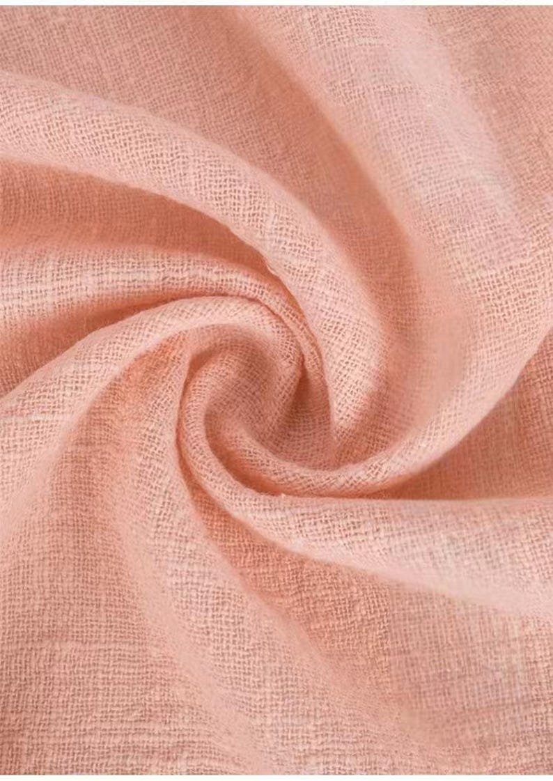 May include: A soft, light peach-colored fabric with a visible woven texture. The material is gathered into a central swirl, creating deep, flowing folds and shadows that highlight the open, airy weave of the textile across the entire surface.