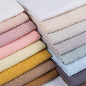 May include: A collection of folded fabric swatches in a variety of colors including white, cream, light pink, dusty rose, pale yellow, mustard yellow, tan, brown, light gray, sage green, sky blue, and dark charcoal, all featuring a textured weave.