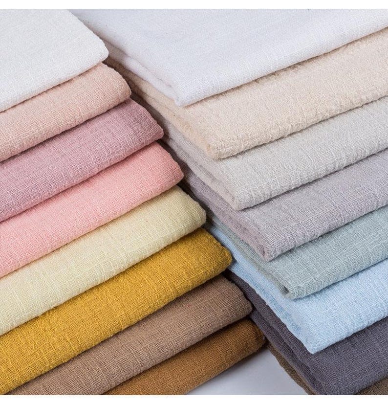 May include: A collection of folded fabric swatches in a variety of colors including white, cream, light pink, dusty rose, pale yellow, mustard yellow, tan, brown, light gray, sage green, sky blue, and dark charcoal, all featuring a textured weave.