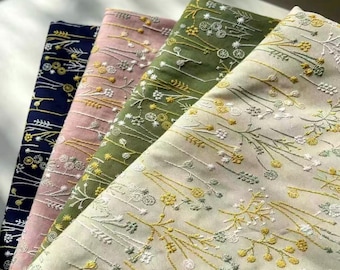 以下が含まれることがあります： 繊細な野草と茎の刺繍パターンが施された、折り畳まれた4枚の布地。布の色はダークネイビー、ソフトピンク、落ち着いたオリーブグリーン、クリーム色で、それぞれ黄色と白の花のステッチで装飾されています。