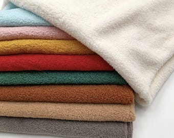 May include: A stack of folded sherpa fleece fabric in various colors including cream, light blue, pink, mustard yellow, red, dark green, brown, tan, and gray. Each piece features a thick, textured, curly pile surface with a soft appearance.