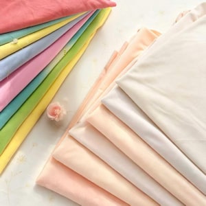 Può includere: Una collezione di tessuti piegati disposti in due pile. La pila a sinistra presenta colori vivaci tra cui rosa, verde acqua, giallo, azzurro e verde. La pila a destra è composta da tonalità neutre color crema, pesca e bianco sporco.