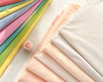 May include: A collection of folded fabric pieces arranged in two stacks. The left stack features vibrant colors including pink, teal, yellow, light blue, and green. The right stack consists of neutral shades of cream, peach, and off-white.