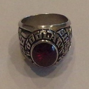 May include: A silver-toned metal class ring featuring a large, round, deep red stone set in the center. The band is embossed with raised lettering that includes the word WOODROW and the number 1997, with additional text visible along the sides.