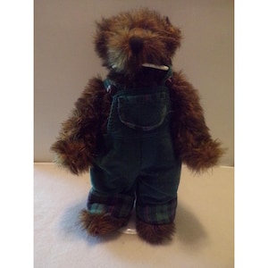 May include: A plush teddy bear with shaggy brown fur wearing dark green corduroy overalls. The overalls feature a front chest pocket and cuffs with a green, blue, and red plaid pattern. A small white tag is visible near the bear's neck area.