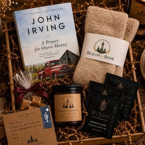 May include: A gift box containing a copy of the book A Prayer for Owen Meany by John Irving, a pair of beige fuzzy socks with a Beacon & Book label, a jar of Pumpkin Orchard candle, three Rishi Masala Chai tea bags, and a bag of wrapped caramels.