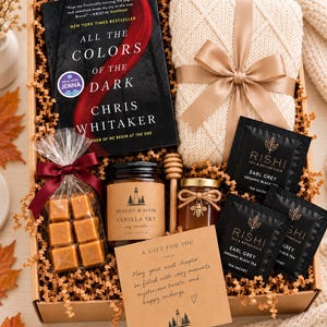 May include: A gift box containing the book All the Colors of the Dark by Chris Whitaker, a cream-colored knit item with a tan ribbon, three Rishi Earl Grey tea sachets, a Beacon & Book vanilla soy candle, honey with a bee charm, and caramels.
