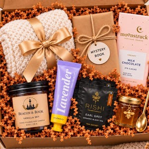 May include: A gift box containing a beige knit item with a gold ribbon, a brown paper-wrapped mystery book, a pink Moonstruck milk chocolate bar, a Beacon & Book vanilla soy candle, a lavender hand cream, Rishi Earl Grey tea, honey, and a gold spoon.