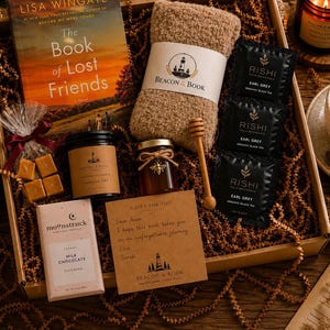 May include: A gift box containing The Book of Lost Friends by Lisa Wingate, a tan fuzzy pair of socks, three Rishi Earl Grey tea bags, a wooden honey dipper, a jar of honey, a candle, a Moonstruck milk chocolate bar, and caramel candies.