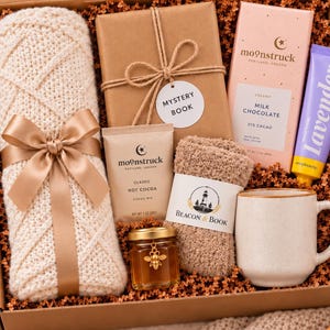 May include: A gift box containing a cream-colored knit blanket with a gold ribbon, a brown paper-wrapped mystery book, a Moonstruck milk chocolate bar, a lavender hand cream, hot cocoa mix, a small honey jar, fuzzy socks, and a mug.