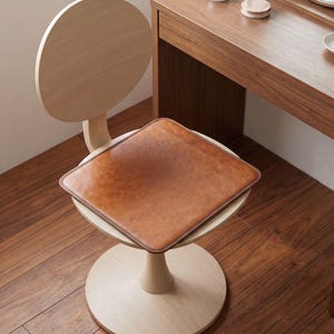 May include: A square, tan leather seat cushion with rounded corners and stitched edges, placed on a light-colored wooden chair with a circular backrest and a pedestal base. The cushion features a smooth, matte finish and a uniform brown tone.