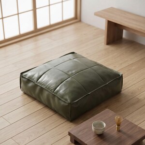 May include: A square floor cushion upholstered in olive green faux leather with a grid-stitched tufted pattern. The cushion features a low-profile design with piped edges, providing a firm and structured appearance for seating on the floor.