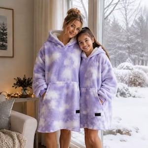 May include: Two oversized hooded sweatshirts made of soft, plush, lavender and white tie-dye fabric. Each garment features a large front pocket, a hood, and a small black rectangular label on the lower front section, lined with white fleece.