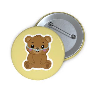 May include: A round pinback button with a pale yellow background featuring a brown cartoon bear wearing a light blue nasal cannula. The bear has dark eyes, a small nose, and pink cheeks. The back of the button is silver with a metal pin mechanism.