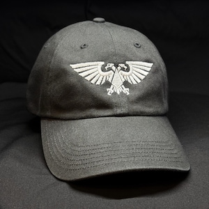 Puede incluir: Una gorra de béisbol gris carbón con un emblema bordado en blanco de un águila bicéfala en el panel frontal. La gorra tiene una visera curva con costuras visibles y una corona estructurada con ojales para la ventilación.