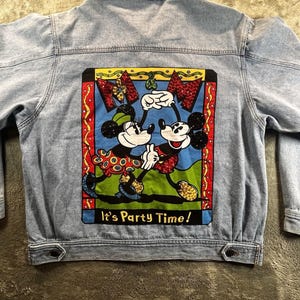 May include: A light blue denim jacket featuring a large, colorful graphic on the back. The print depicts two cartoon mice dancing, surrounded by a red, yellow, and blue border. The text "It's Party Time!" is printed in yellow at the bottom.