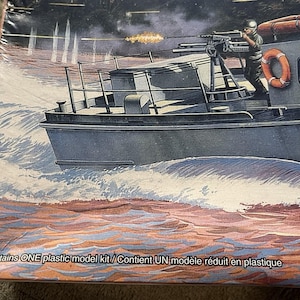 May include: A plastic model kit box featuring an illustration of a grey military patrol boat moving through water. The boat has a mounted machine gun, a person in uniform, and an orange life ring. Text reads: Contains ONE plastic model kit.