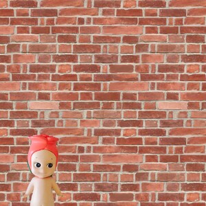 Puede incluir: Una pequeña muñeca de color carne con un sombrero de capullo de rosa roja se encuentra frente a una pared de ladrillo rojo.