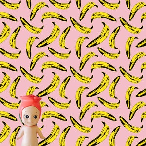 May include: A pink background with a repeating pattern of yellow banana shapes. A small doll with a pink rose head is standing in the centre of the image.
