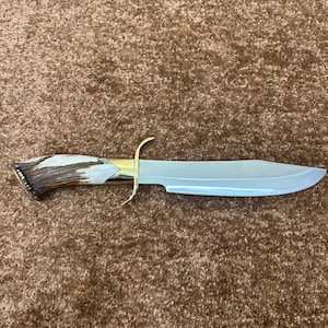 Puede incluir: Un cuchillo de hoja fija con una hoja de acero pulido en tono plateado, una guarda de metal en tono dorado y un mango de asta texturizado en tonos marrón y blanco. El cuchillo presenta un diseño curvo con una punta afilada.