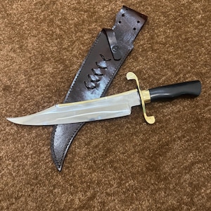 May include: A fixed-blade knife with a polished silver-tone steel blade, a smooth black handle, and a brass crossguard. The knife is accompanied by a dark brown leather sheath featuring decorative black lacing and a snap-button closure strap.