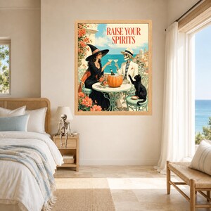 May include: A framed wall art print titled RAISE YOUR SPIRITS featuring a witch in a black dress and a skeleton in a white suit toasting with glasses at a table. A black cat sits on a chair next to a pumpkin, with a coastal Mediterranean backdrop.