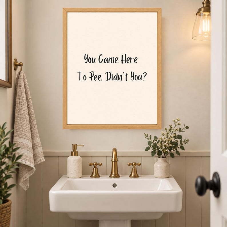 May include: A rectangular wall art piece in a light wood frame featuring a cream-colored background with the black text "You Came Here To Pee, Didn't You?" centered on the print. The artwork is displayed above a white bathroom sink basin.