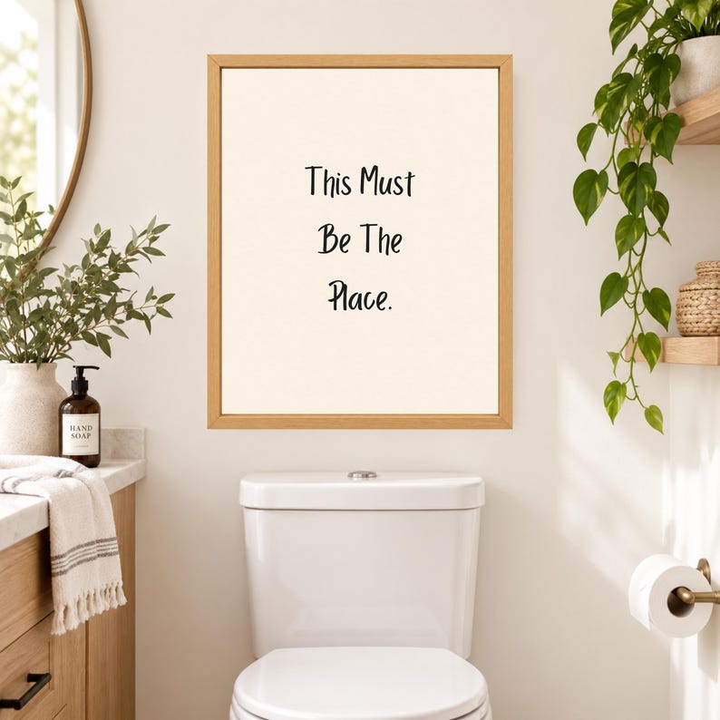May include: A rectangular wall art print in a light wood frame featuring the black text "This Must Be The Place." on a cream-colored background. The print is displayed on a plain wall above a white toilet in a bathroom setting.