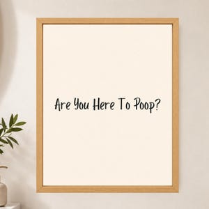 May include: A framed wall art piece featuring a light cream-colored background with the black text "Are You Here To Poop?" centered in the middle. The artwork is enclosed in a simple, light-toned wooden frame with a clean, rectangular border.