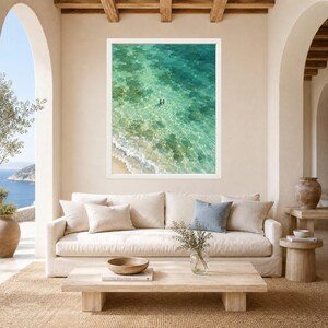 May include: A framed wall art print featuring an aerial view of turquoise ocean water with a sandy shoreline and two small figures swimming in the clear sea. The artwork is displayed in a white frame hanging on a beige wall above a beige sofa.