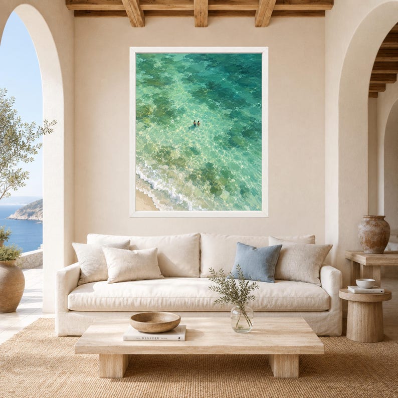 May include: A framed wall art print featuring an aerial view of turquoise ocean water with a sandy shoreline and two small figures swimming in the clear sea. The artwork is displayed in a white frame hanging on a beige wall above a beige sofa.