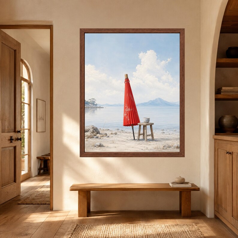 May include: A framed wall art print featuring a coastal scene with a bright red closed parasol standing upright on a sandy beach next to a small wooden stool, with calm blue water, distant mountains, and a light blue sky with white clouds.