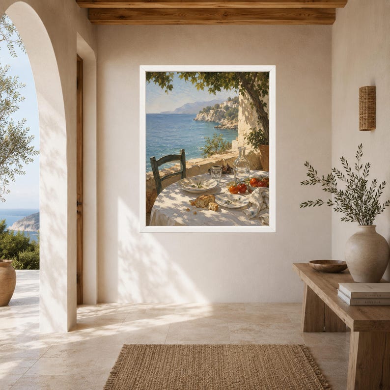 May include: A framed wall art print depicting a Mediterranean coastal scene with a table set for a meal under a tree. The table features a white cloth, plates, a glass carafe, and red tomatoes, overlooking a blue sea and rocky cliffs in the distance.