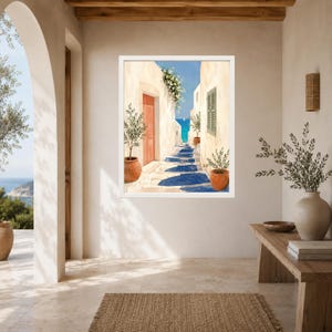 May include: A framed wall art print depicting a narrow Mediterranean alleyway with white stucco walls, a bright orange door, green shutters, and potted olive trees leading toward a vibrant blue sea under a clear sky with deep blue shadows.