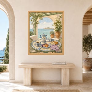 May include: A framed wall art print depicting a Mediterranean terrace scene with a round tiled table, a blue and white teapot, cups, fresh fruit, and bread. The background shows a coastal view with a pergola, potted plants, and the sea.