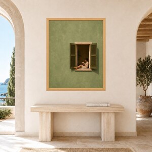 May include: A framed wall art print featuring a sage green wall with an open window and dark green shutters. A person sits on the window ledge reading a book. The artwork is displayed on a light-colored wall above a rectangular stone console table.